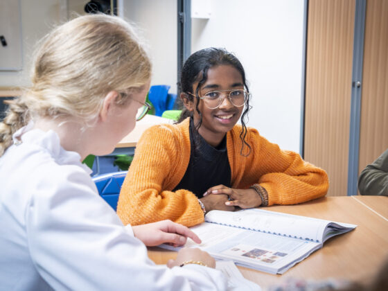 Leerling, kijkend in de camera, is bezig met schoolwerk en leert samen met een andere leerling uit een boek